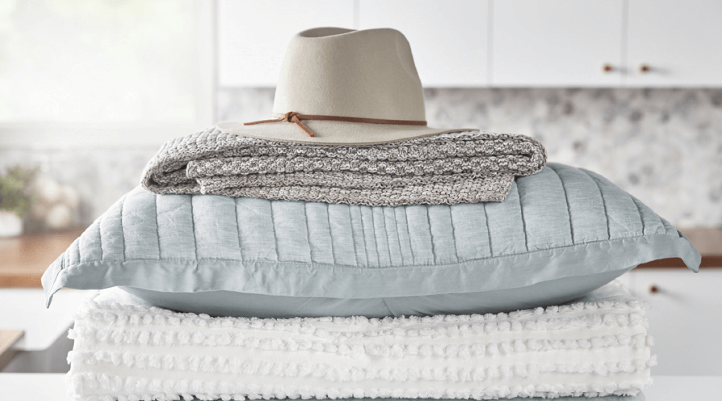 A stack of blankets, pillows and a hat sit on a laundry room counter.