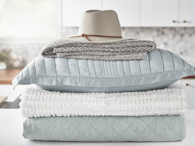 A stack of blankets, pillows and a hat sit on a laundry room counter.